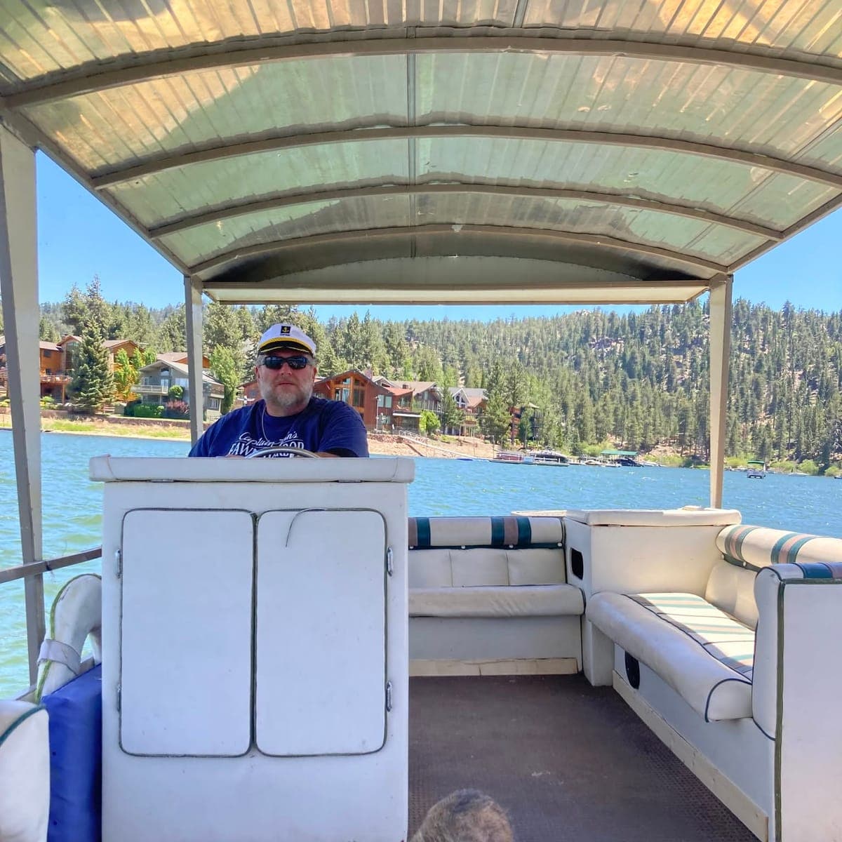 Captain Ben at the wheel of the covered tour boat, lakeside cabins and forest along the shore behind him.